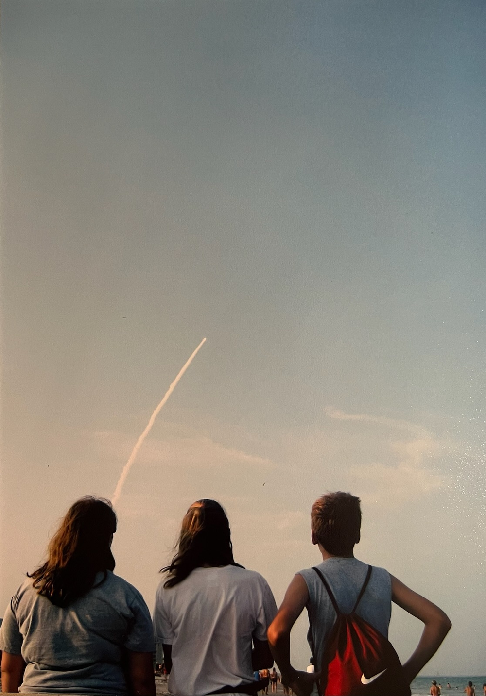 Three people watch a shuttle launch from the beach.